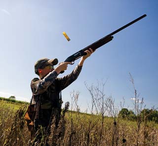 A hunter in a field fires a shotgun. The shell is ejected from the firearm.