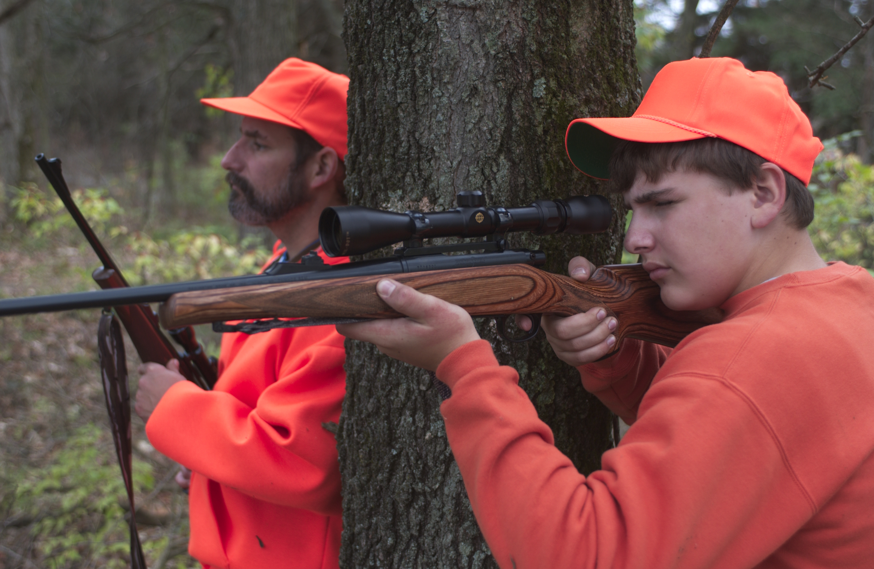 Two hunters in safety orange sit by a tree. One is taking aim at a deer.